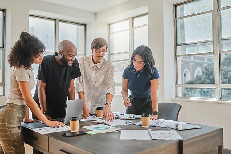 Three women and a man in an office stand as they review a series of documents spread across a table.