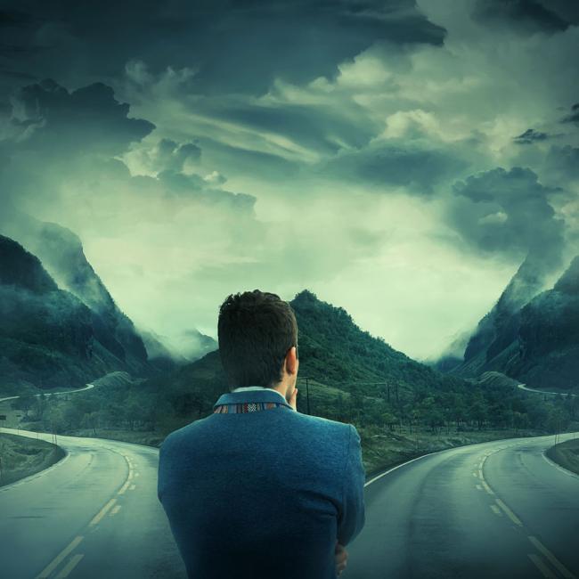 Man stands with his back facing us while two roads diverge in front of him.