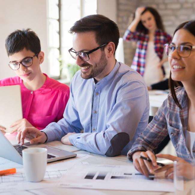 Three people in foreground wearing glasses looking a a shared screen. A woman stands in the background with one hand on her head and the other on her hip.